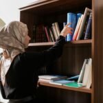 A woman in a headscarf reaches for a book on a wooden bookshelf in a serene indoor setting.